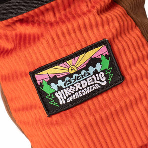 A patch featuring a pink and yellow sunset amongst mountains and trees with psychi logo affixed to a orange and brown corduroy rock climbing chalk bag