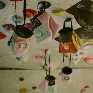 A collection of corduroy climbing chalk bags and bouldering buckets in orange, pink, mint green, and brown hanging from holds on a climbing wall at Parthian Climbing Centre Manchester