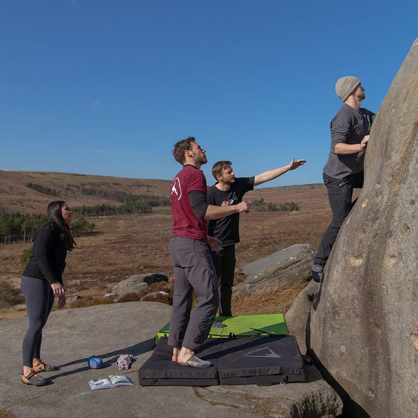 Two climbing instructors supervise as a beginner boulderer climbs up a boulder at Stanage Plantation in the Peak District.