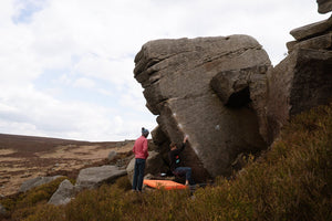 A climber clinging to a boulder in moorland environment while his friend stands watching. 