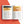 An open printed recipe book laid flat on a white background with large orange-red geometric shapes behind it. The spread shows two pages with food photos, headings, and blocks of text, including a banana and nuts in a bowl on one page and a baked good on the other.