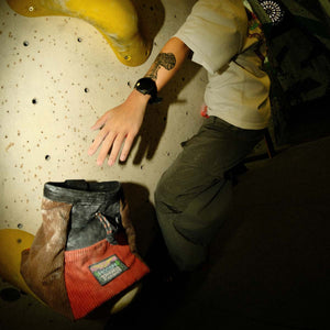 A boulderer hanging onto a climbing wall reaches down towards an orange and brown psychi chalk bucket.