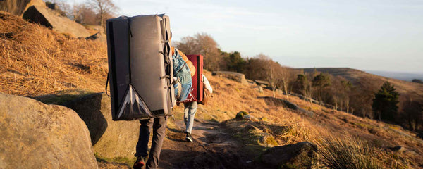 Two climbers walking along a path, amongst large boulders, carrying bouldering pads at Stanage Edge in the Peak District