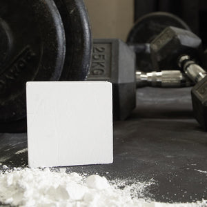 A white chalk block sitting on a gym matt in front of some dumbbells with chalk powder in front of it.