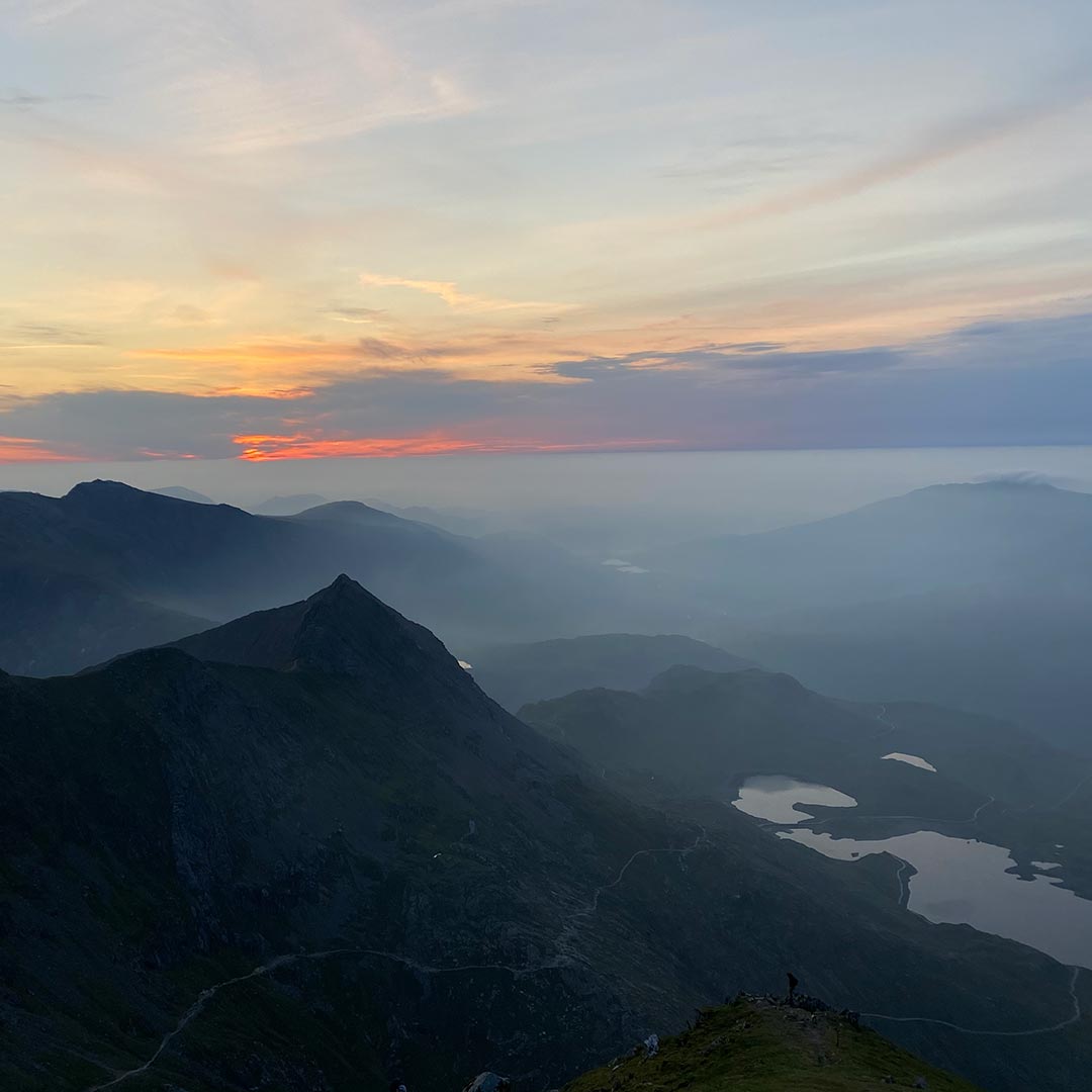 The sun sets behind a series of misty mountains. A sharp peak in the foreground gives way to a lake in the Lake District 