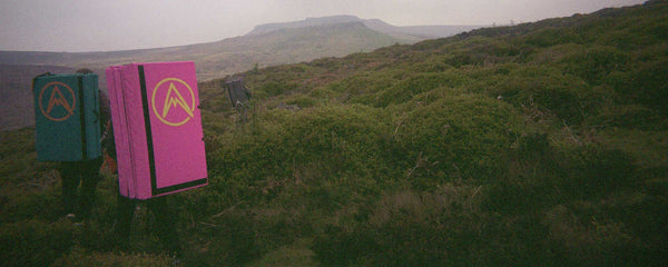 Three climbers carrying bouldering pads walk on a path overgrown with heather at Burbage Edge in the Peak District. 