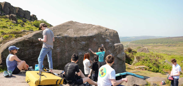 A group of climbers gathered around a large grit stone boulder in the peak district, with a green moorland vista in the background.