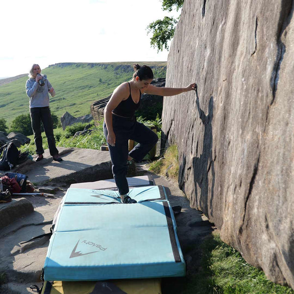 A female boulderer stands on a blue bouldering pad and prepares to climb a rock face at stanage plantation in the peak district