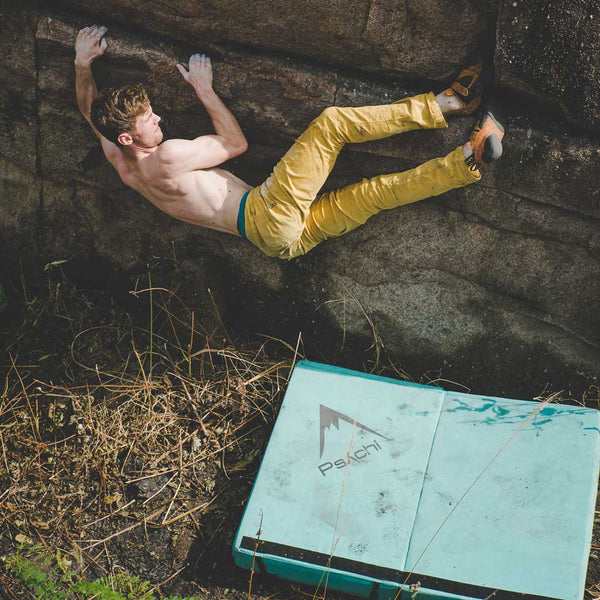 A male boulderer clings to a boulder face above a bright blue bouldering safety mat in Burbage