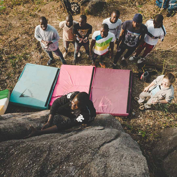 A boulderer climbing a crack in a large boulder in the Ivory Coast. Below him are nine other climbers and three bouldering crash pades