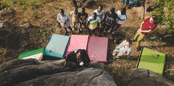 A group of locals in a village in the Ivory Coast watch their friend climb a boulder for the first time. He clings to the rock above five different coloured psychi bouldering pads. 