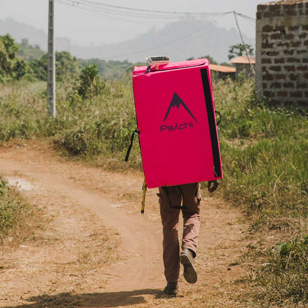 A boulderer walks along a dusty path carrying a pink bouldering crash pad in the Ivory Coast