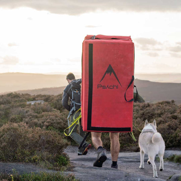 two rock climbers and a dog walk along a rocky path in stanage, one is carrying a thick red bouldering crash pad